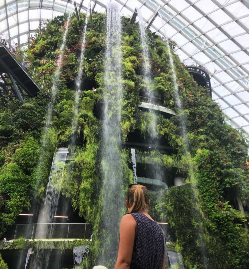Travel blogger standing in front of an indoor waterfall covered in greenery inside the cloud conservatory