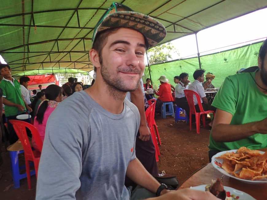 Travel blogger Tj smiling and eating at a table full of different Burmese dishes served family style at a Burmese wedding in a village
