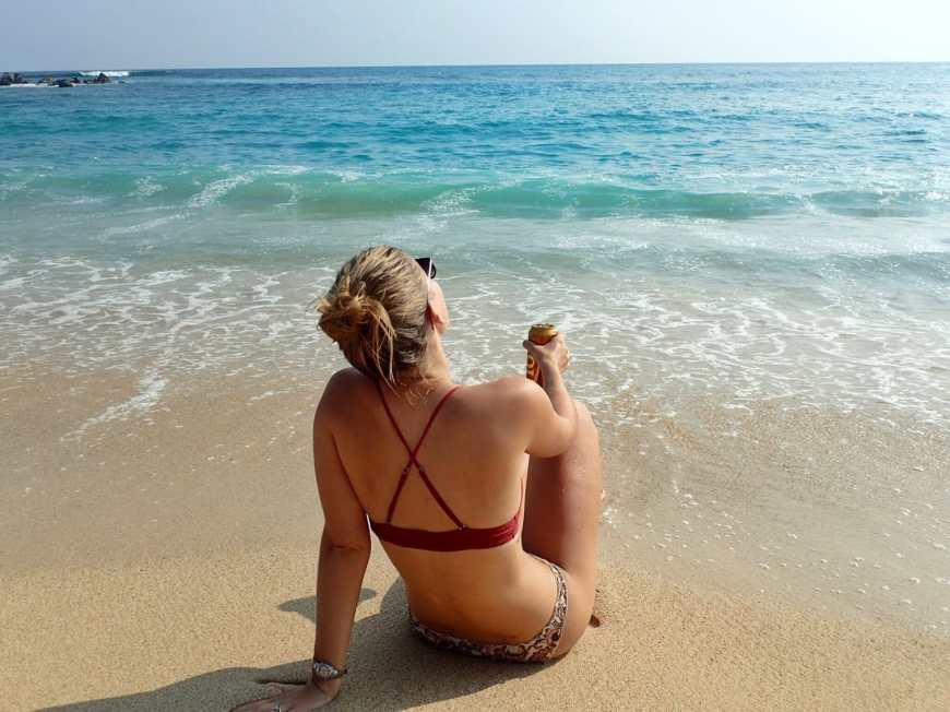 Woman sitting on the beach in a bikini facing the ocean as waves crash over her toes and she holds a lion beer