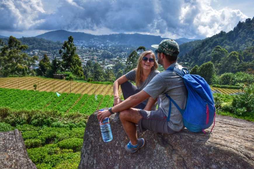 Travel and adventure couple laughing as they sit on an enormous boulder overlooking tea fields and the town of Nuwara Eliya in Sri Lanka