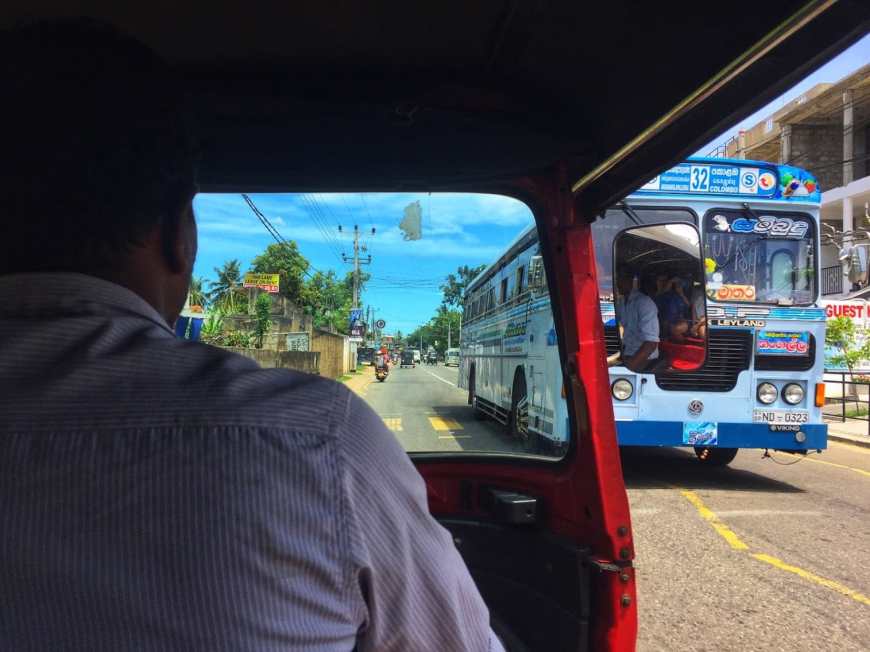 Image shot from the inside of a red tuk tuk looking forward to the driver in a button down as we drive past a large blue buss
