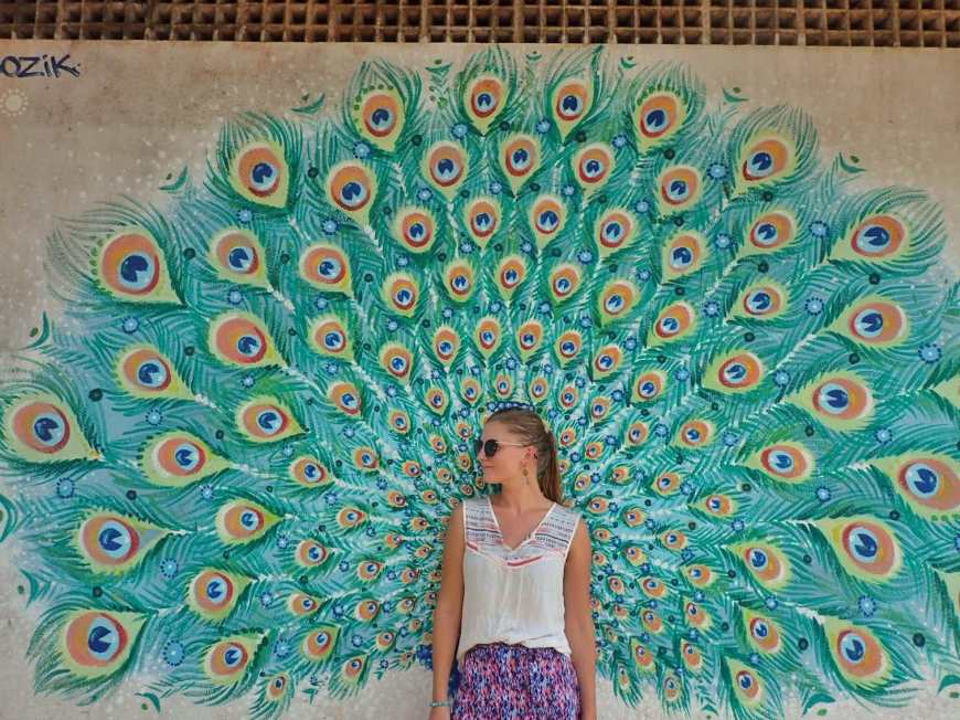 Woman standing in front of a peacock mural as the feathers span out behind her like wings